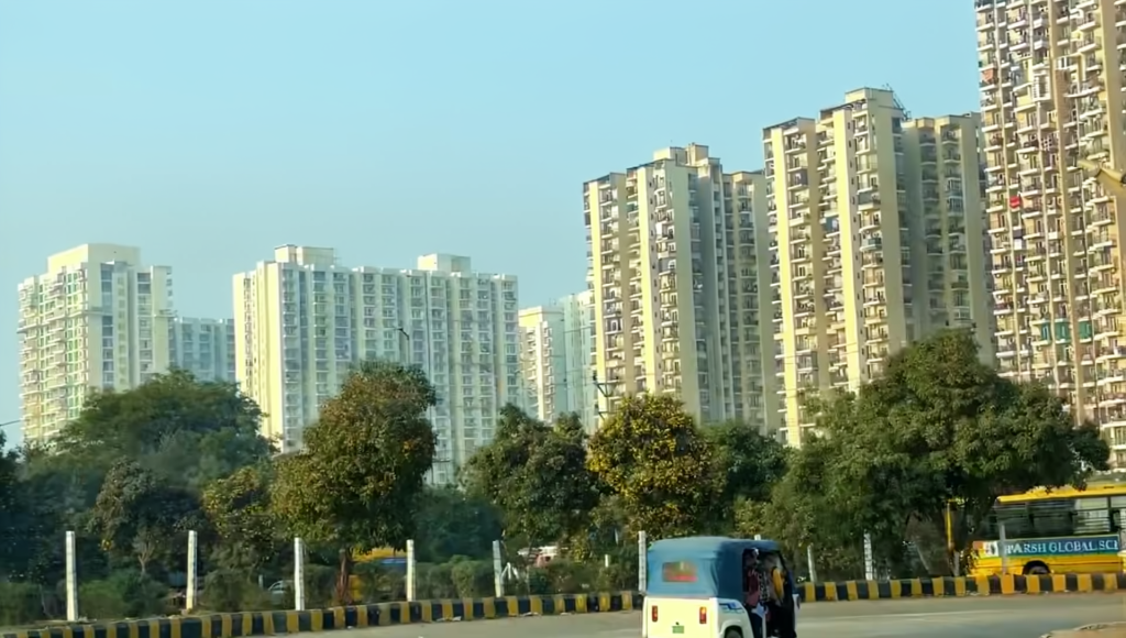 Wide tree-lined road in Noida with modern residential societies in the background
